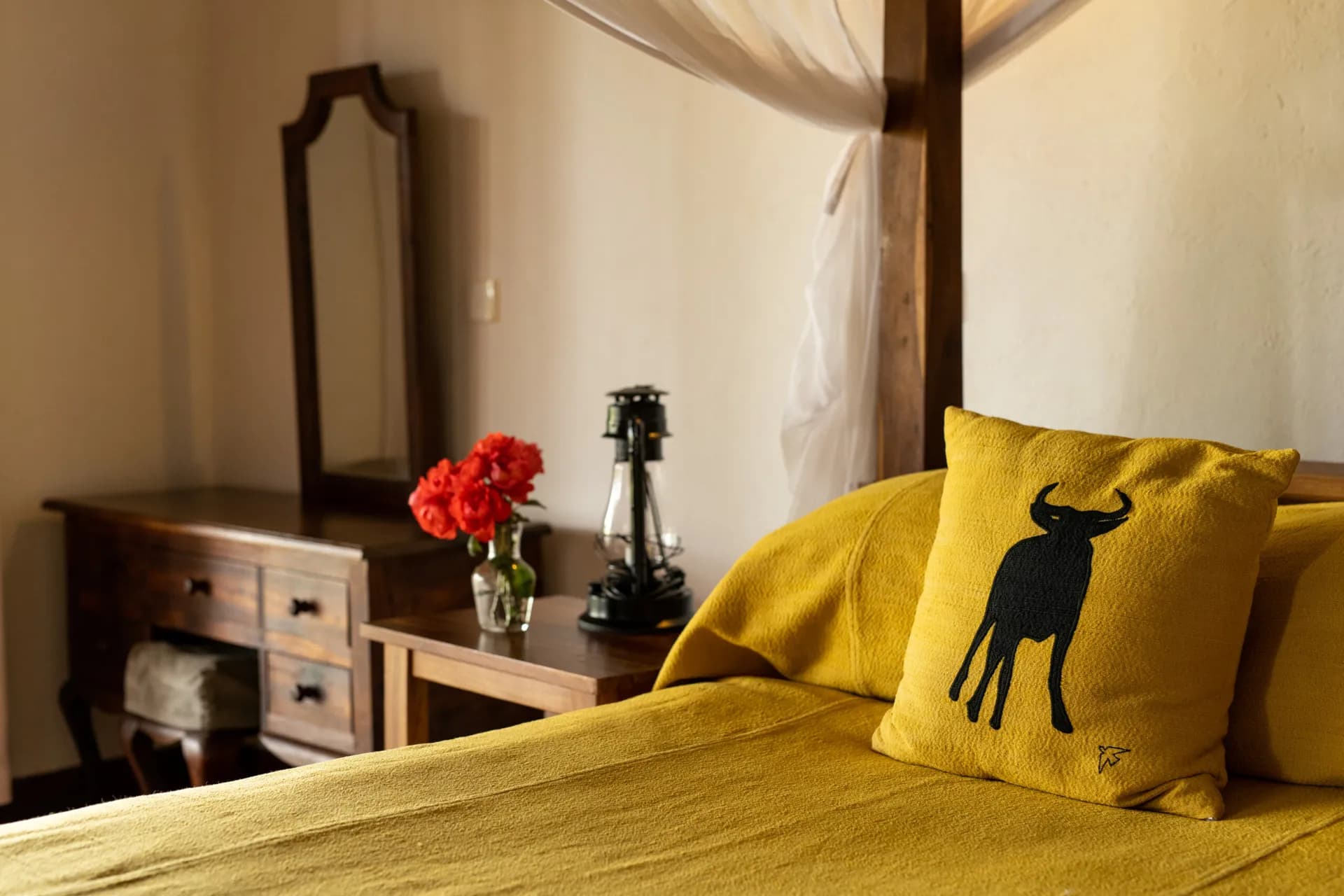 Yellow wildebeest cushion, red roses and a lantern beside a four-poster bed at Ngorongoro Farm House
