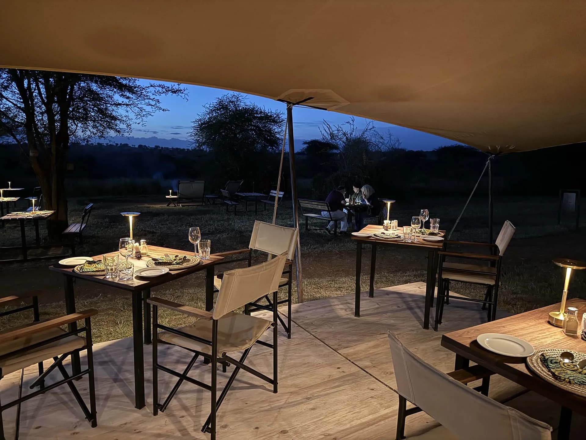 Lamp-lit dinner tables under canvas at dusk at The Whisper Tarangire, with acacia trees beyond